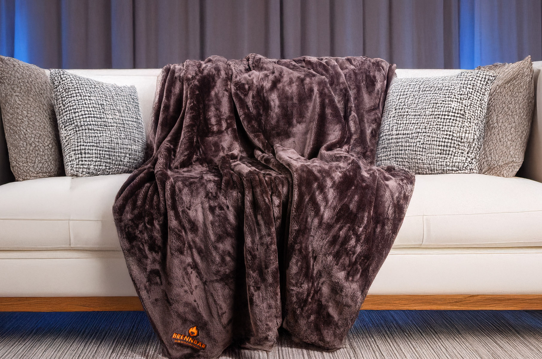 Kuscheldecke in Schoko-Farbe mit Brennbär-Logo liegt ausgebreitet auf weißem Sofa mit grauen und braunen Kissen