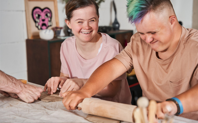 Zwei Menschen mit Trisomie 21 lachen und arbeiten gemeinsam beim Ausrollen von Teig