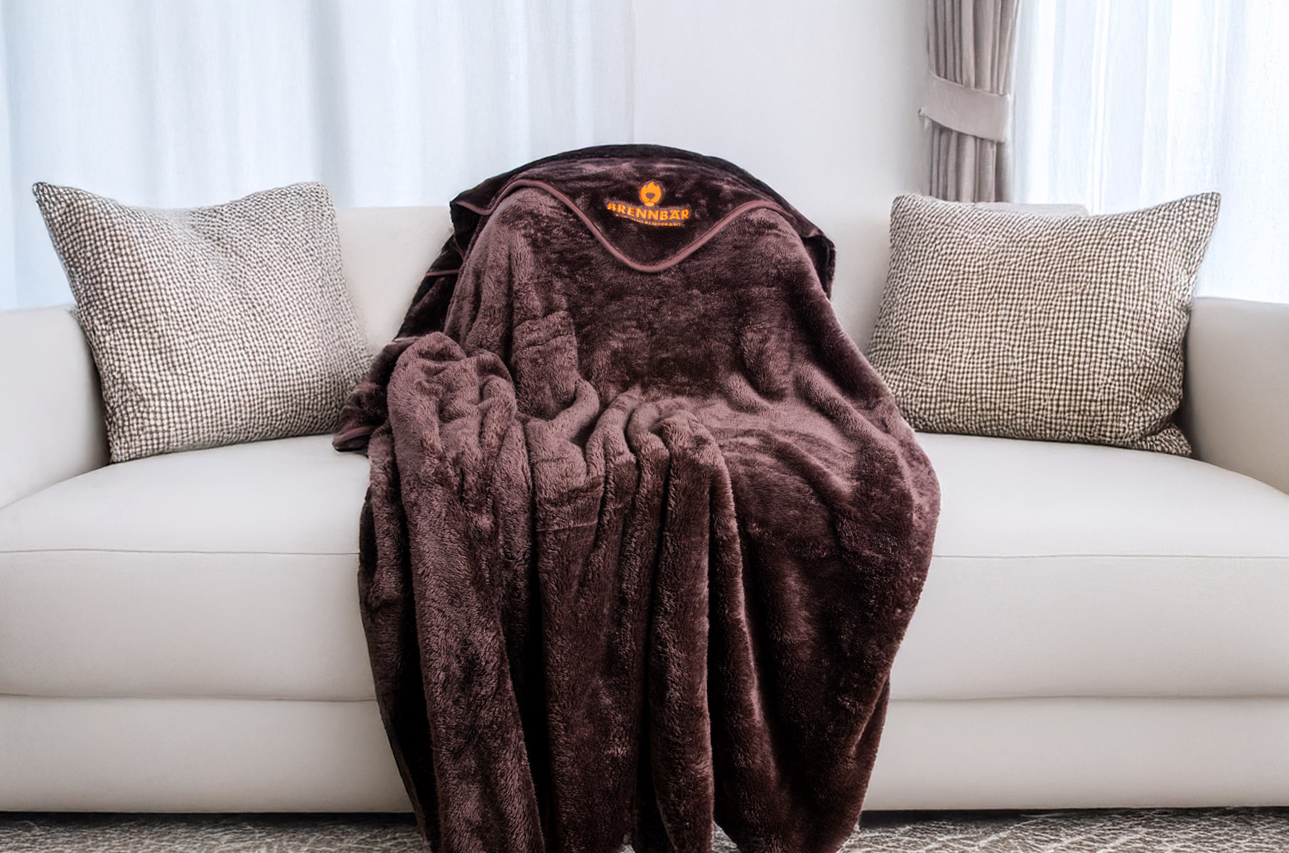 Kuscheldecke in Schoko-Farbe mit Brennbär-Logo liegt ausgebreitet auf weißem Sofa mit grauen Kissen